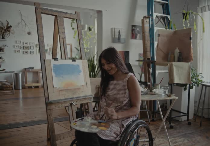 Lady on a wheelchair talking while holding a brush and palette inside an art studio at midday