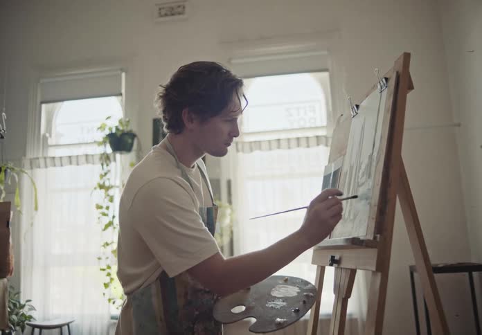 A painter man is painting with a brush a picture and he is holding a drawing board with paint