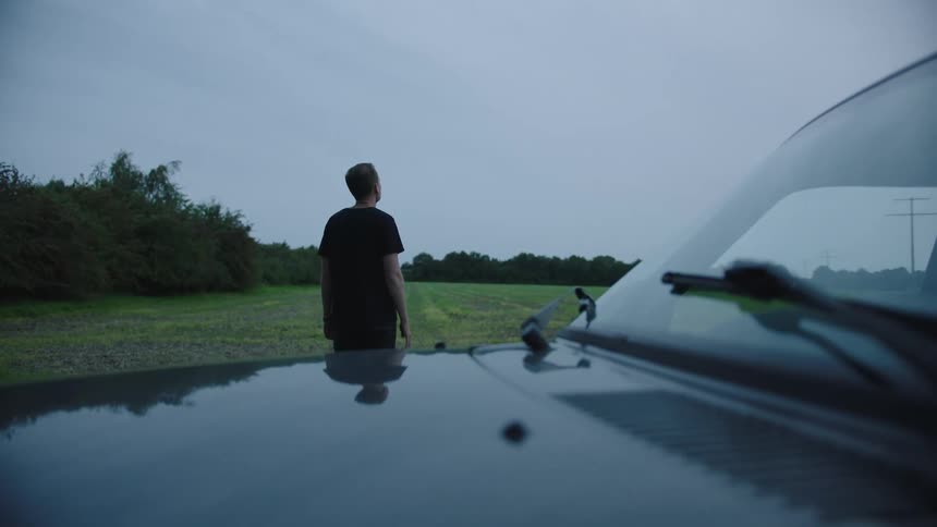 View of a man standing outside the car on a lawn watching the cloudy day