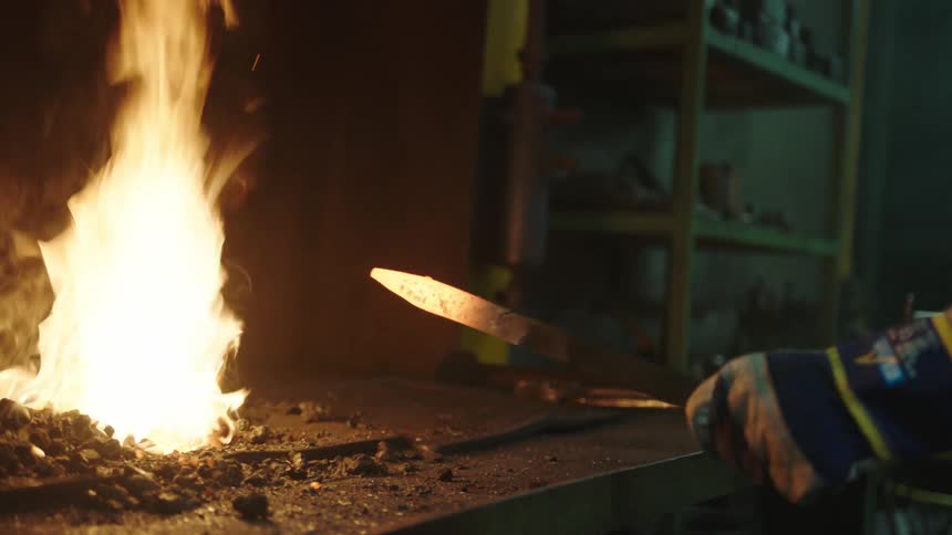 Closeup of a mans arm grabbing a hot iron from the fire and forging it with a hammer