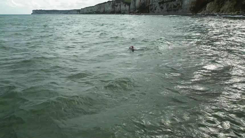 An adult swims in the ocean on a partly cloudy day with cliffs in the background