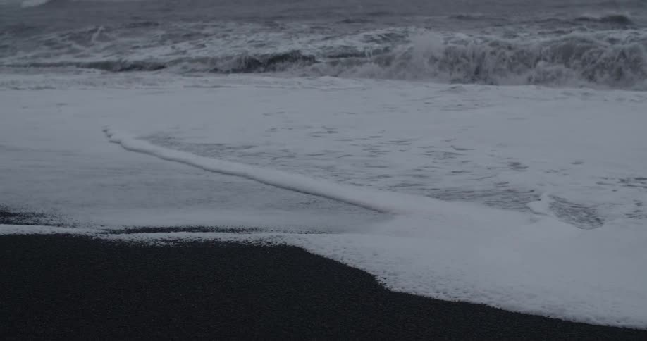 Foam covers black sand as a wave retreats from the shore and another comes in