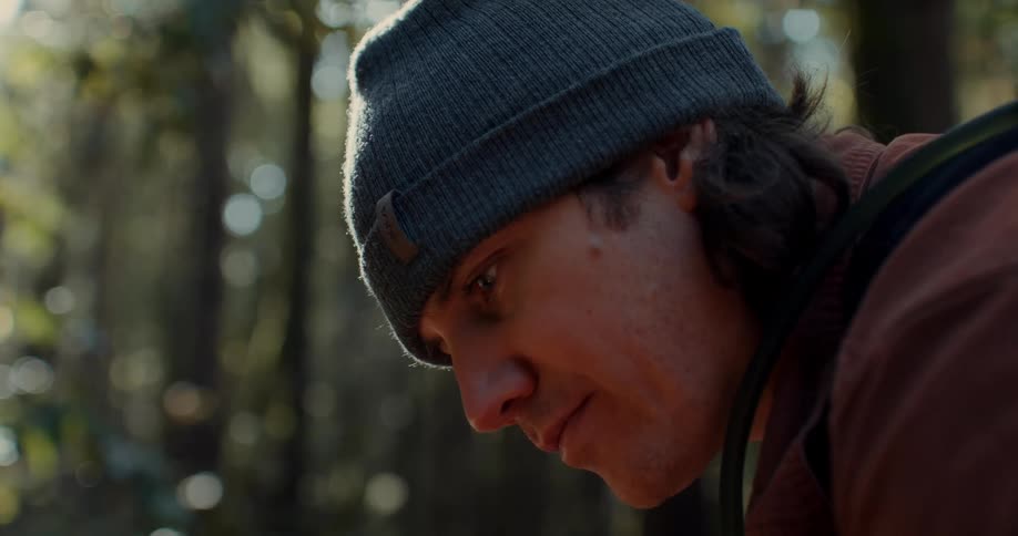 A man wearing a beanie who is looking down intently and behind him are some trees