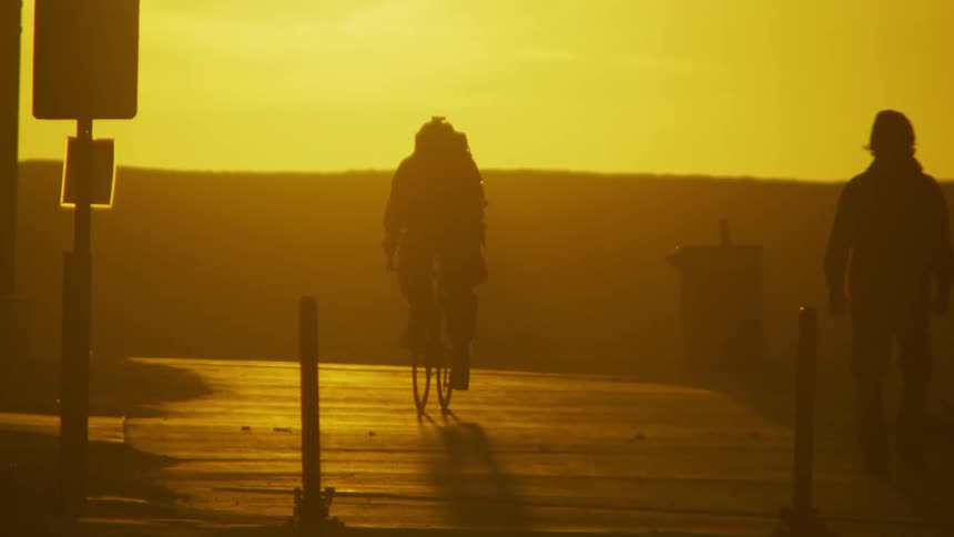 Cyclist and pedestrian move along trail silhouetted against sunlight during sunset