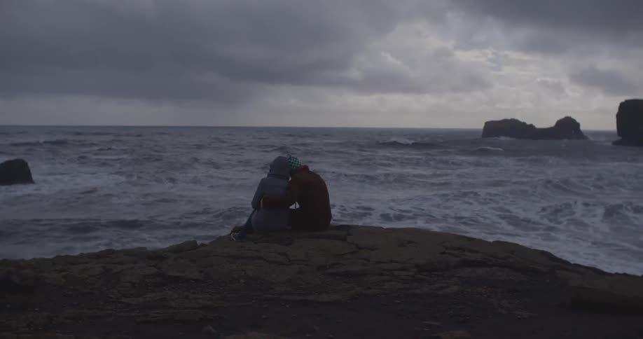 A man has his arms around a woman as they sit on a rock on a shoreline