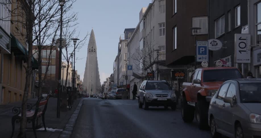 A person crosses a city street with many vehicles parked on the side