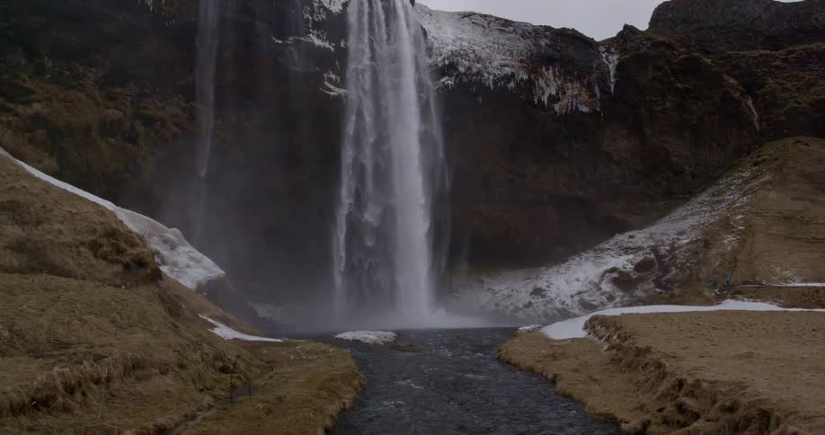 A large waterfall in slow motion flows into a river below with a dusting of snow on the scene