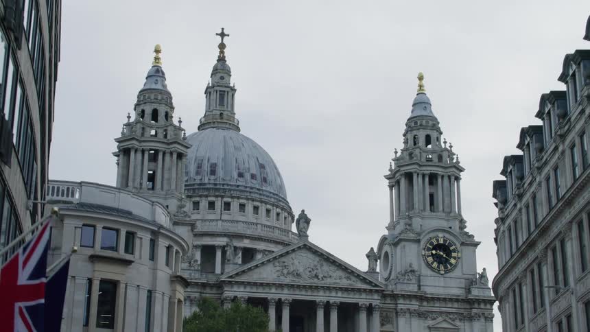 Airplane flies over landmark buildings including a clock tower and a domed cathedral on an overcast day