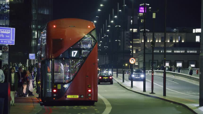 Red double decker bus stops on city street near pedestrians at night