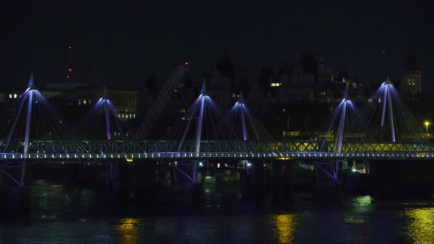 Crane towers over bridge crossing reflective river water near buildings at night