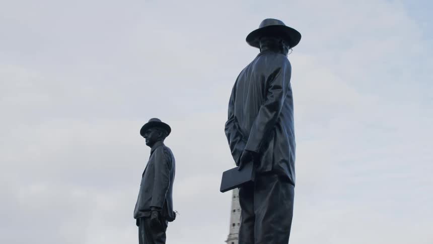 Sculpture of two men facing in opposite directions stands near domed government building