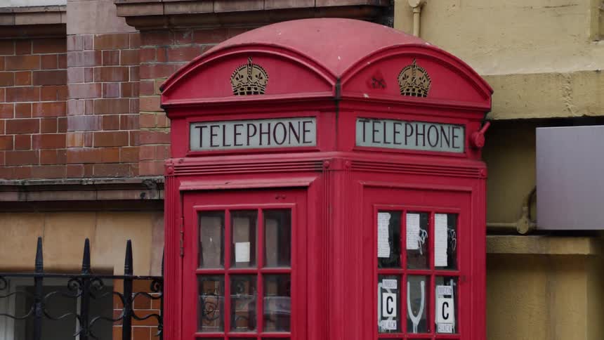 Pedestrians pass red public phone booth situated near building and iron fence
