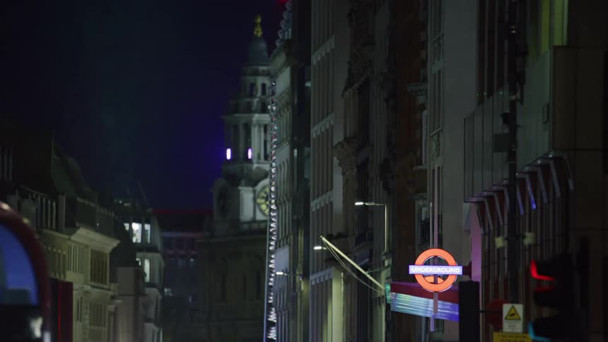 Red double decker bus drives by buildings and london underground sign at night