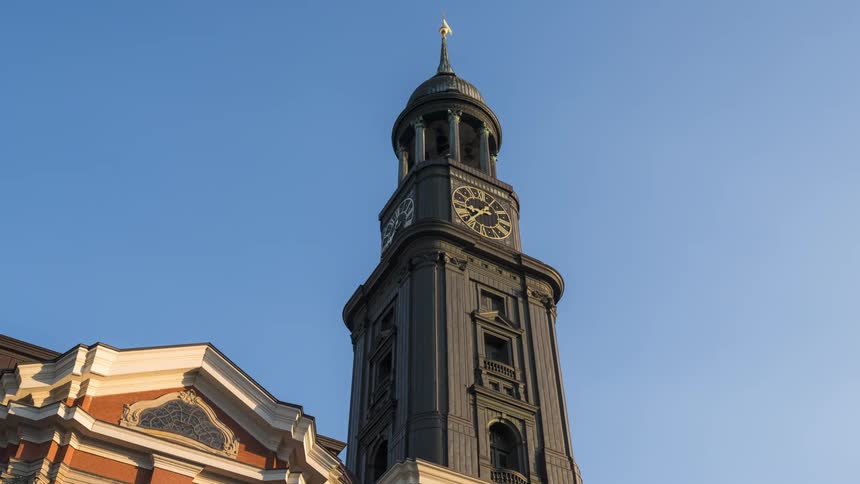 Dark colored clock tower stands over tree and reddish and tan building on a windy day