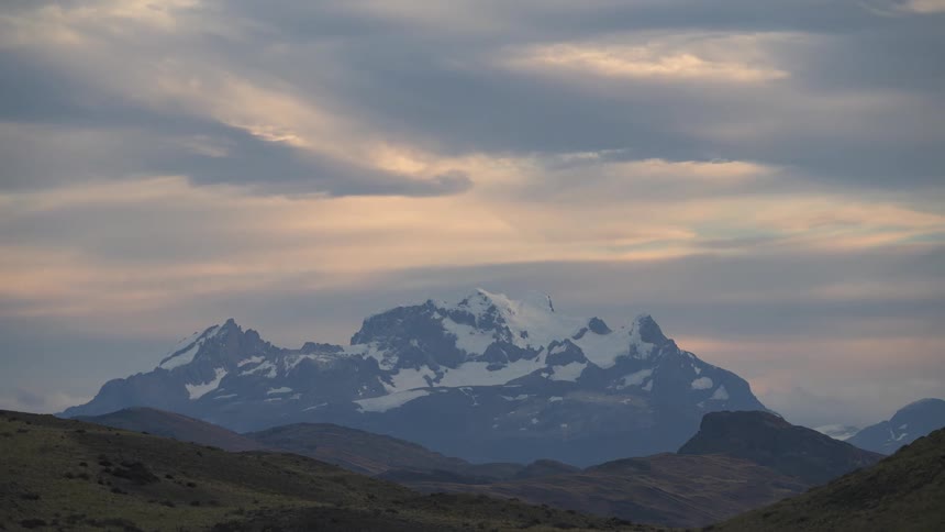Clouds darken while passing over tall snowy mountain towering over hills
