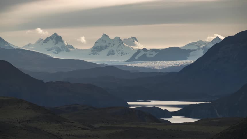Clouds flow over glacier filled plateau towering over lake in mountain range
