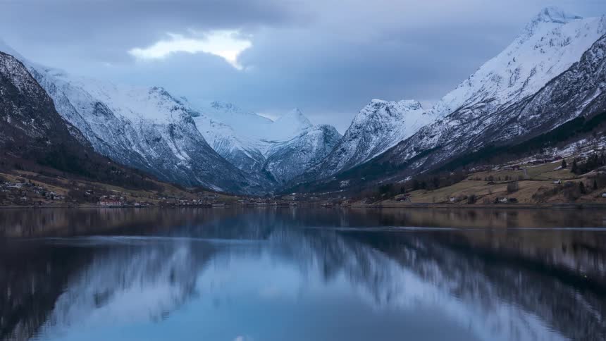 Lake water reflects mountains surrounding rural town on a cloudy day