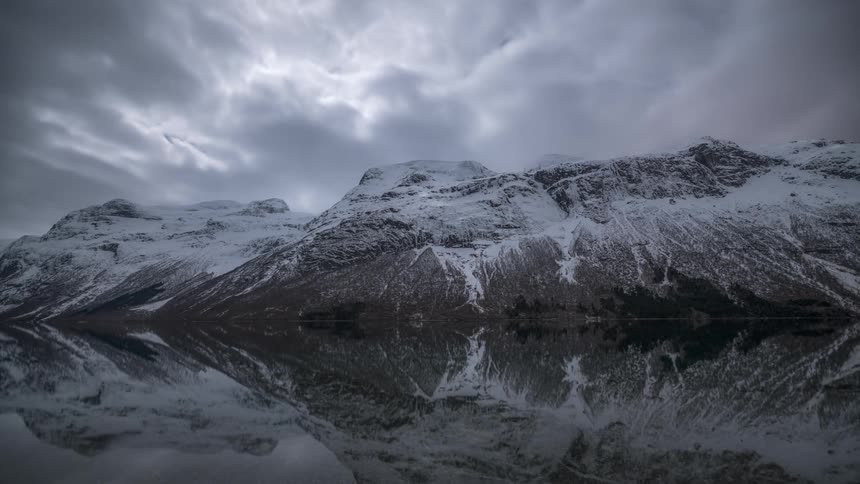 A scene of snowcapped mountains and their reflection in a lake under a cloudy sky
