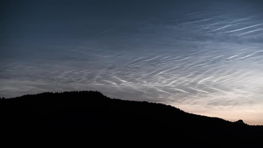 A forested mountain range silhouetted against a bright display of Noctilucent Clouds (NLC's)
