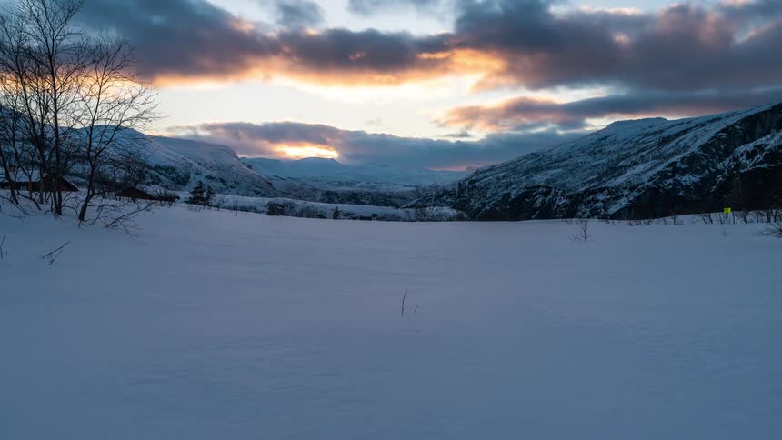 A snowy landscape with bare trees and mountains during sunset