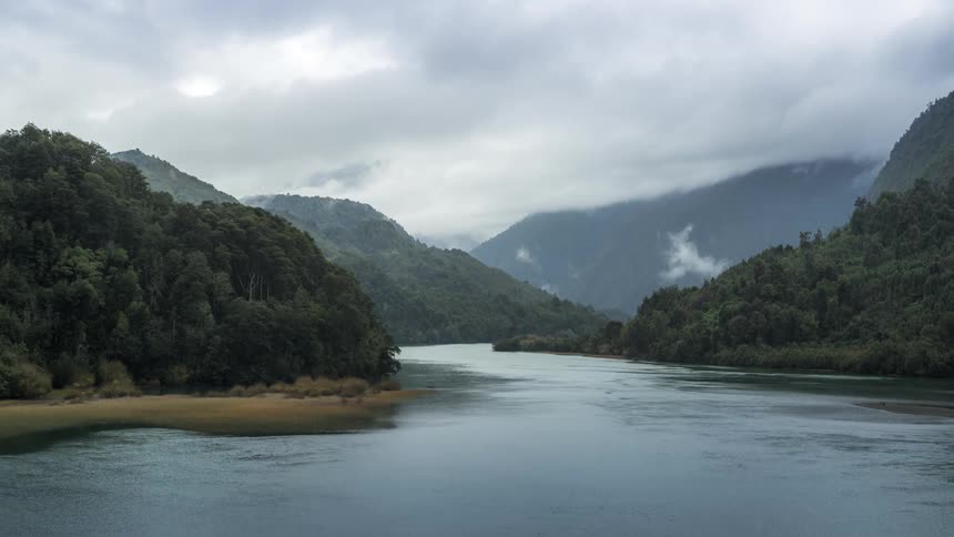 Mountain range with forests and a flowing river in the center with passing clouds on a cloudy day in time lapse