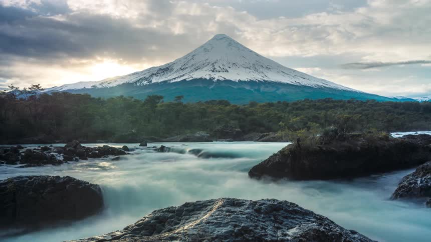 Sun shines over slope of snow topped mountain onto river on a heavily clouded day