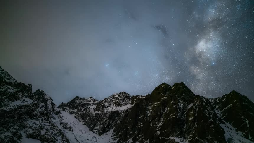 Landscape of a starry sky with nebula with clouds passing in timelapse and mountain ranges covered in snow