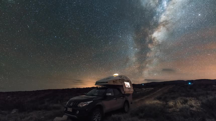 A truck with a camper sits under a sky filled with the milky way