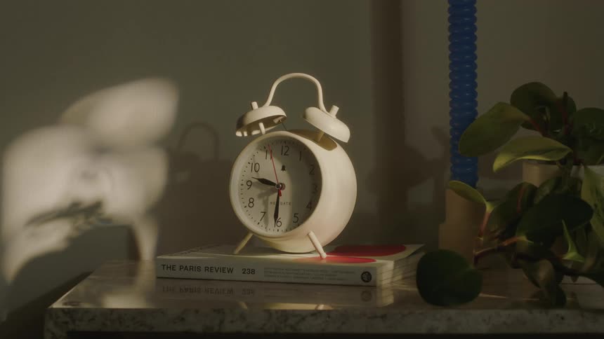 An alarm clock ringing, a woman's hand turns it off and knocks it off the table