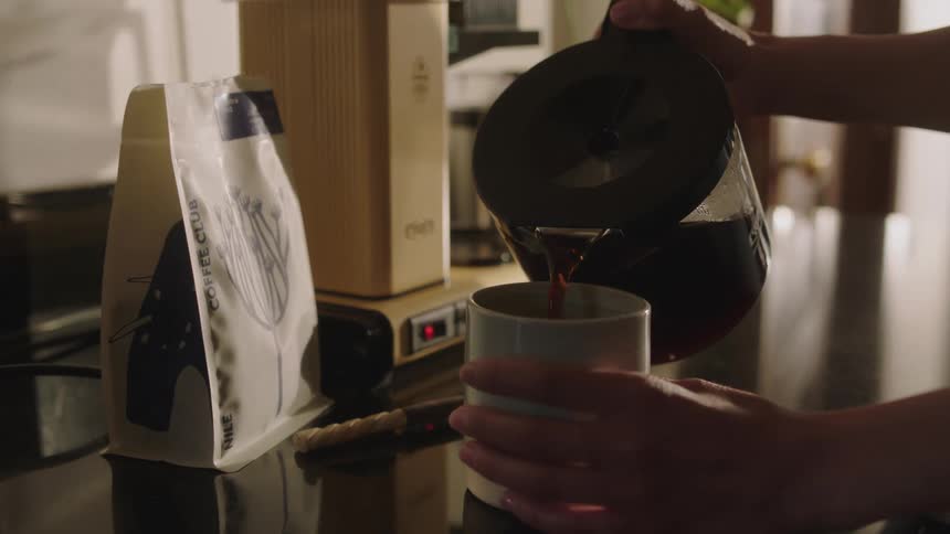 A person pours coffee into a mug in the kitchen with a bag of coffee and Moccmaster coffee machine in the background