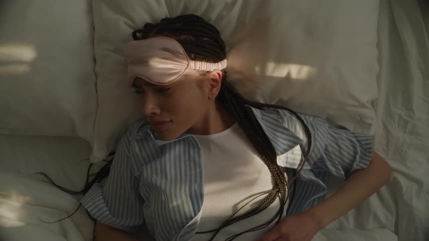 Overhead of a young woman waking up in bed, lifting her sleep mask