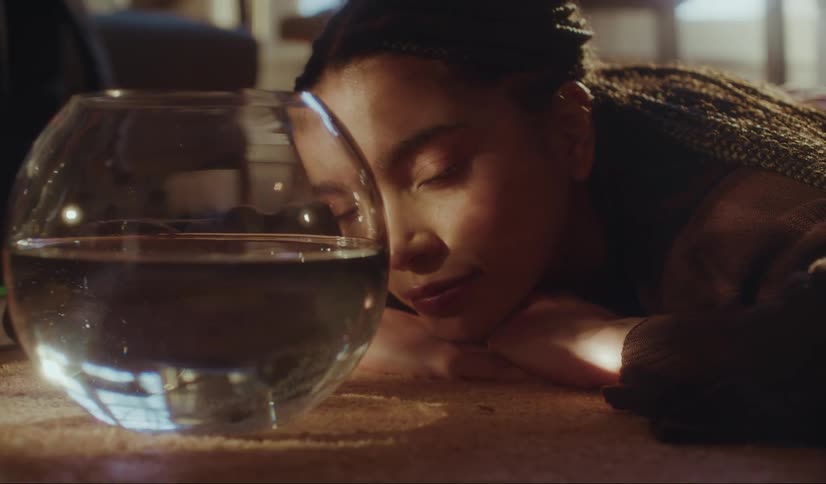 A young woman lays on the floor watching a goldfish swim around a bowl