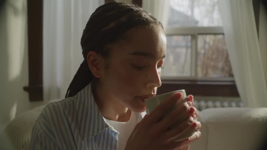 A young woman looking out the window at the morning sunlight sipping a cup of coffee
