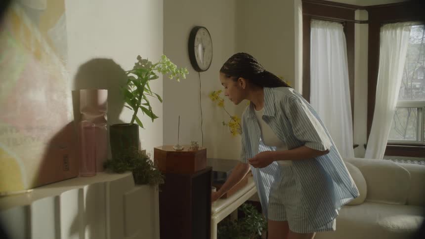 A young woman in pajamas walks into the living room with a cup of coffee and lights an incense