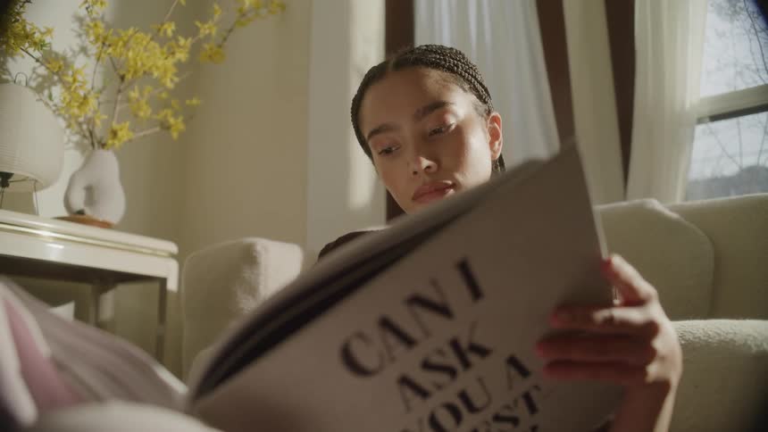 A young woman reading a large book in a bright living room with flowers