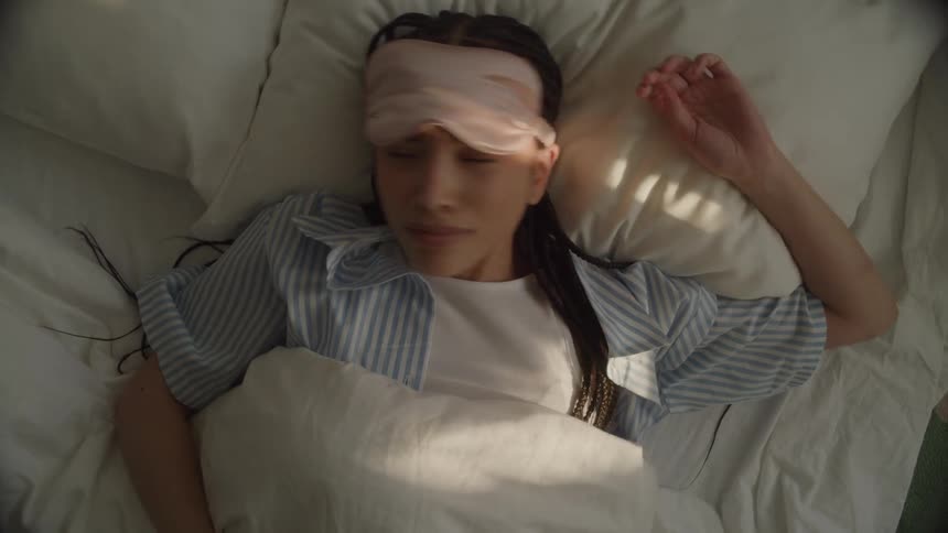 Overhead of a young woman waking up in bed, lifting her sleep mask