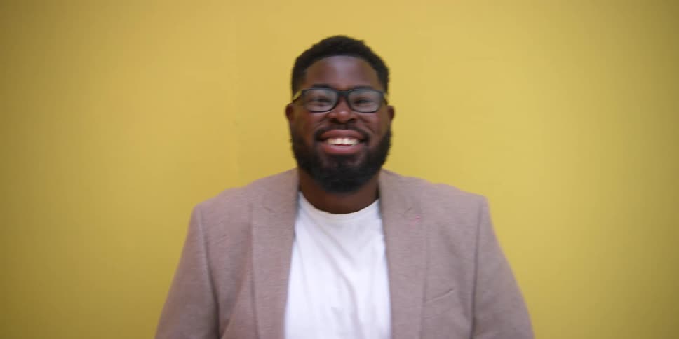 Black male adult in front of a yellow backdrop wearing a suit and glasses is smiling