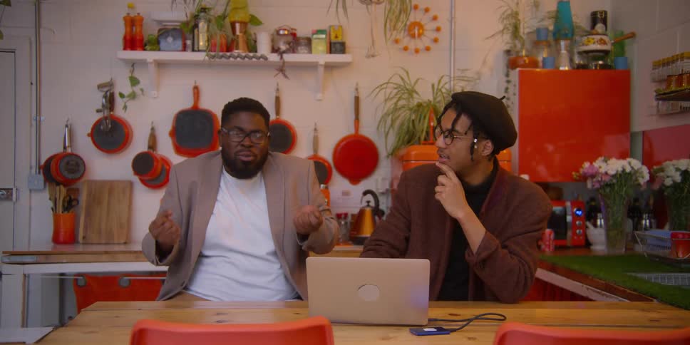A conversation between two individuals at a wooden table with a laptop and orange utensils in a kitchen