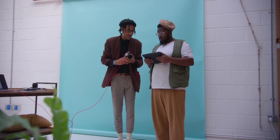 Photogapher takes photos of the model wit a hat beardd glasses a green vest and brown pants in the studio