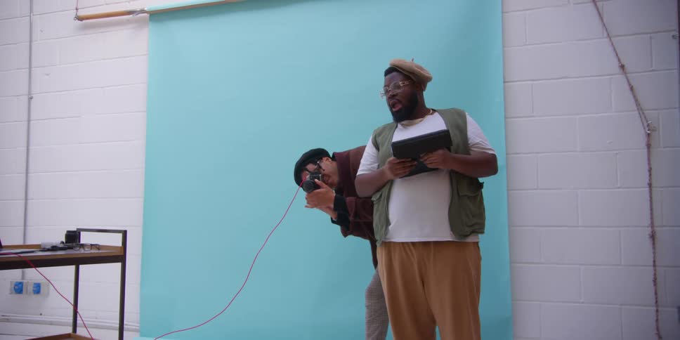 Photographer takes photo of man wearing a hat glasses a green vest white t shirt and brown pants in the studio