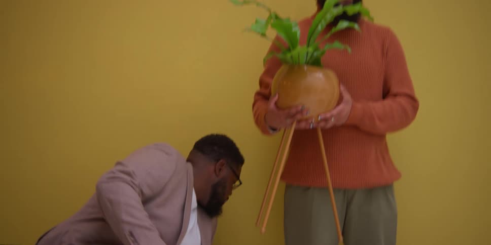 A man posing with a potted plant in his hands while another man fixes his clothes