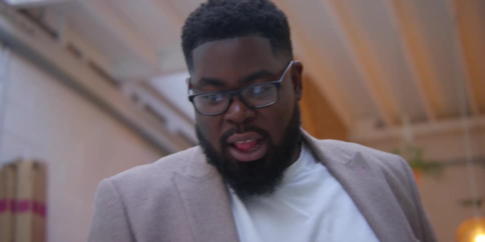 Black male adult standing in doors looking down than up and over as he moves his mouth speaking