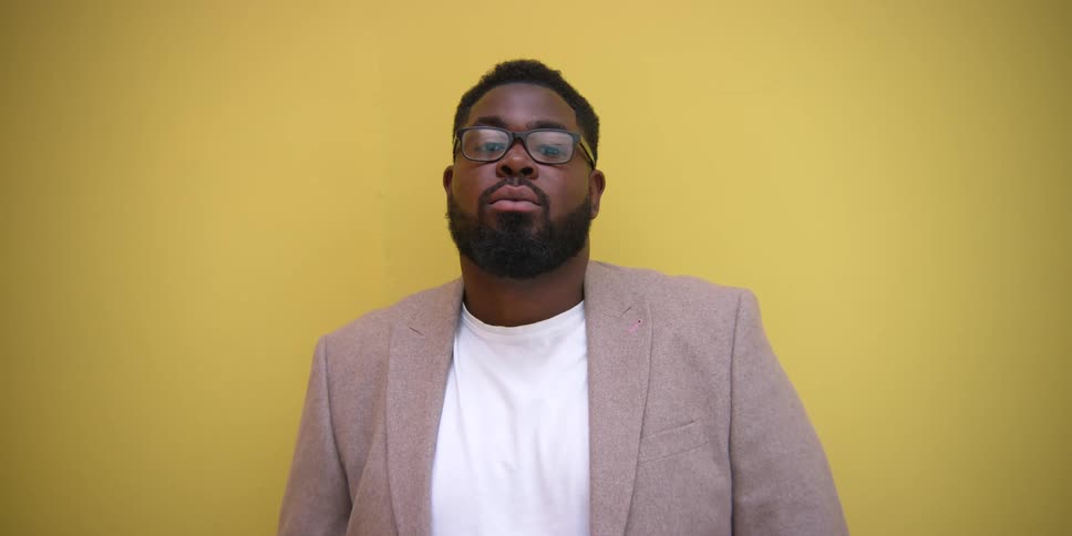 A person standing against a yellow background wearing a white shirt and a lightcolored blazer