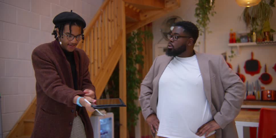 A person in a brown coat and a beret holding a tablet talks with another person in a white shirt and grey blazer standing in a room with wooden stairs and shelving