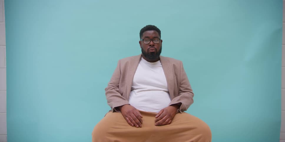 A black man sits in front of a blue background while fussing with his clothes