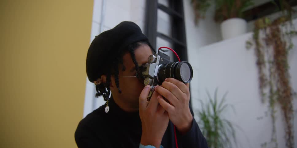 Young black adult male photographer taking pictures with a handheld camera and smiling in an indoor photo studio filled with natural light