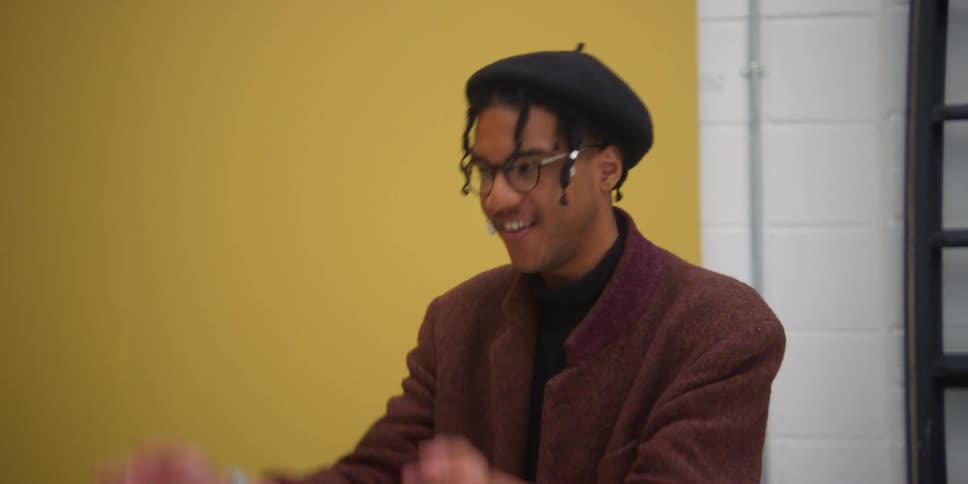 A person gesturing with their hands against a yellow wall wearing a beret and a brown jacket