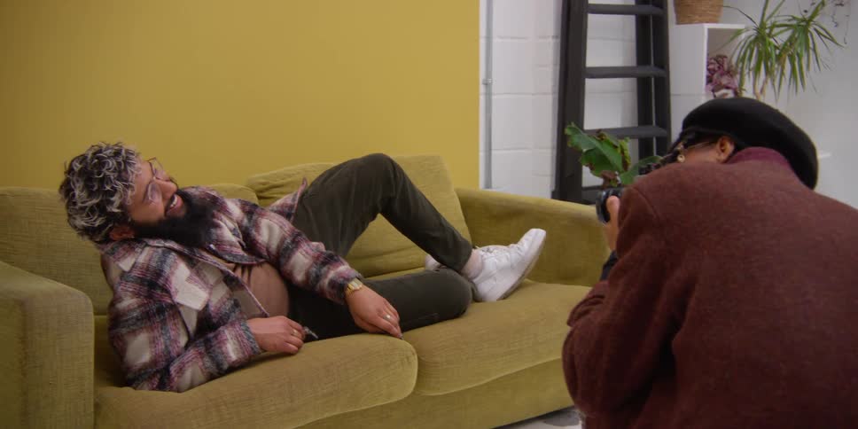 A man lying on a couch is laughing as his pictures are being taken by another man holding a camera