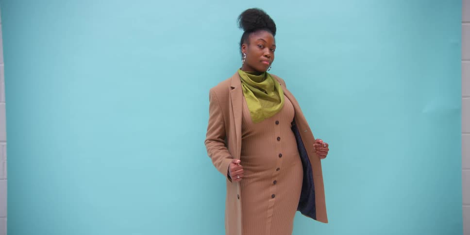 A woman wearing a brown blazer jacket over a dress and a green scarf poses for the camera
