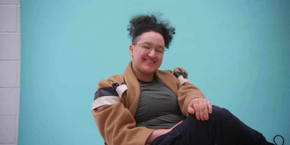 Person of ambiguous gender sitting down on a chair indoors smiling next to a blue wall